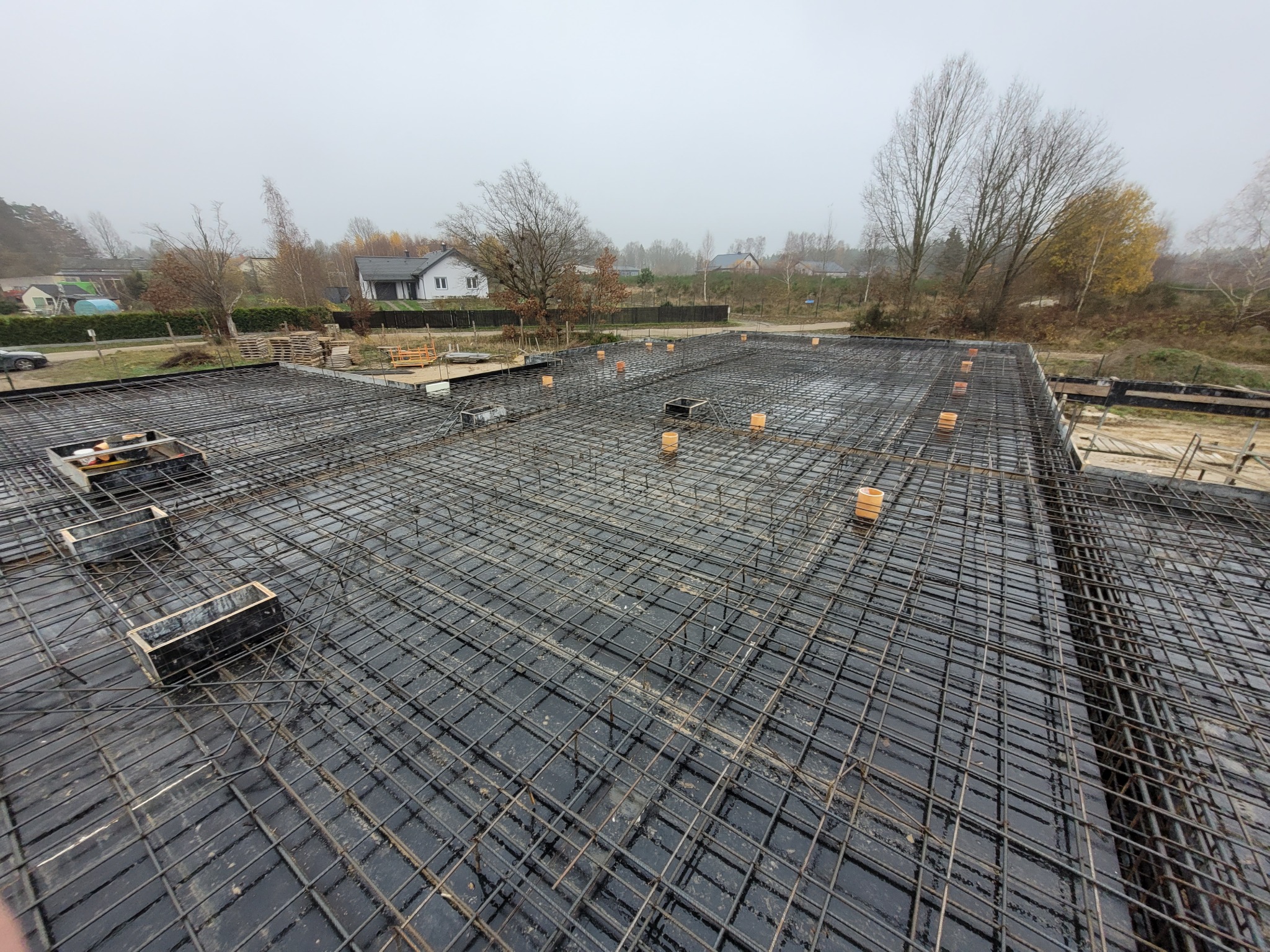 Zbrojenie płyty fundamentowej z widocznymi elementami szalunkowymi i dystansami, na tle zabudowy jednorodzinnej w pochmurny dzień.