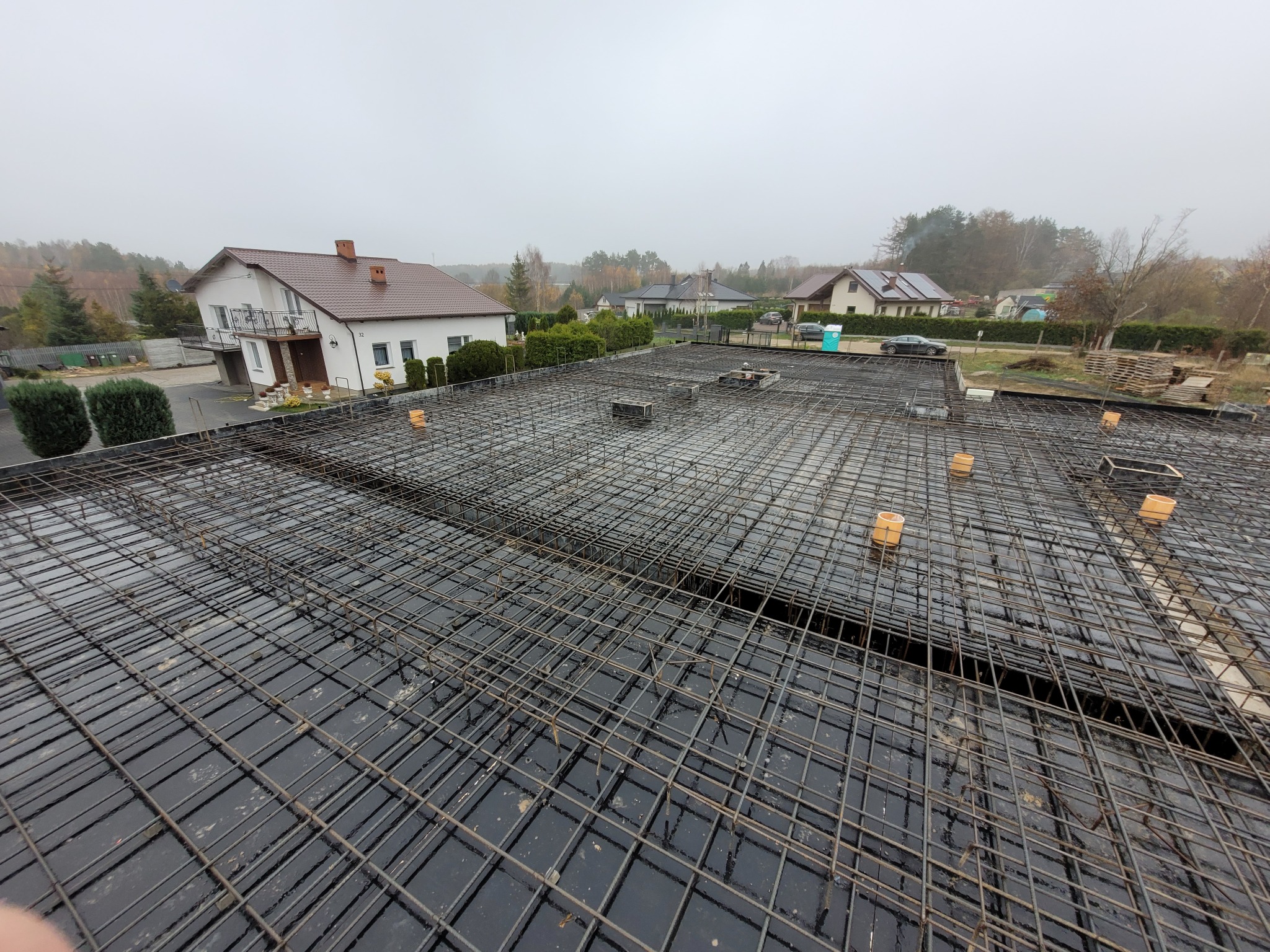 Siatka zbrojeniowa na czarnej folii, przygotowana do zalania betonem, widok z góry na tle zabudowy jednorodzinnej w pochmurny dzień. Konstrukcja fundamentów.