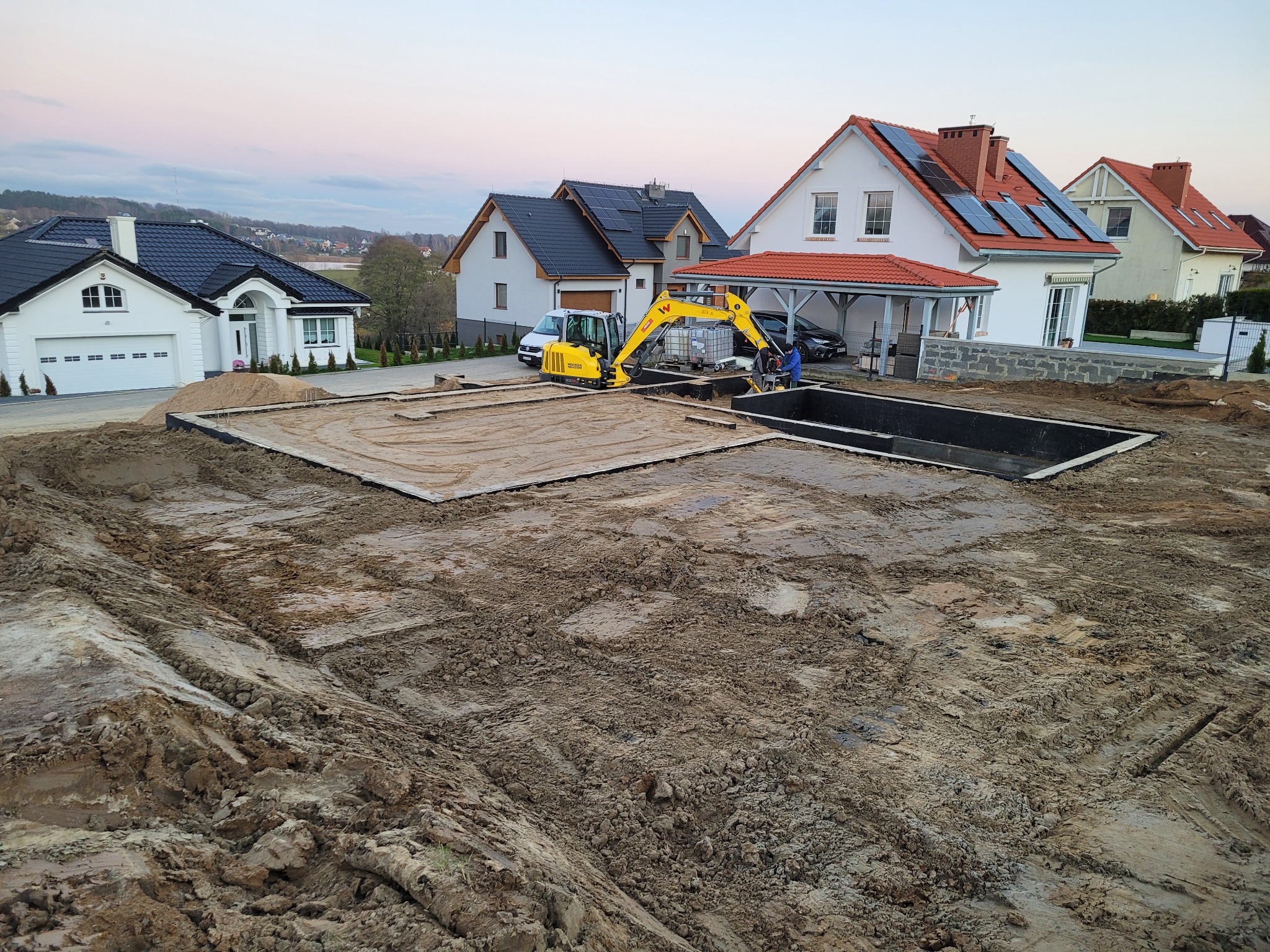 Żółta mini koparka przy budowie fundamentów pod basen na terenie budowy z domami w tle. Widoczne wykopane fundamenty i pracownicy.