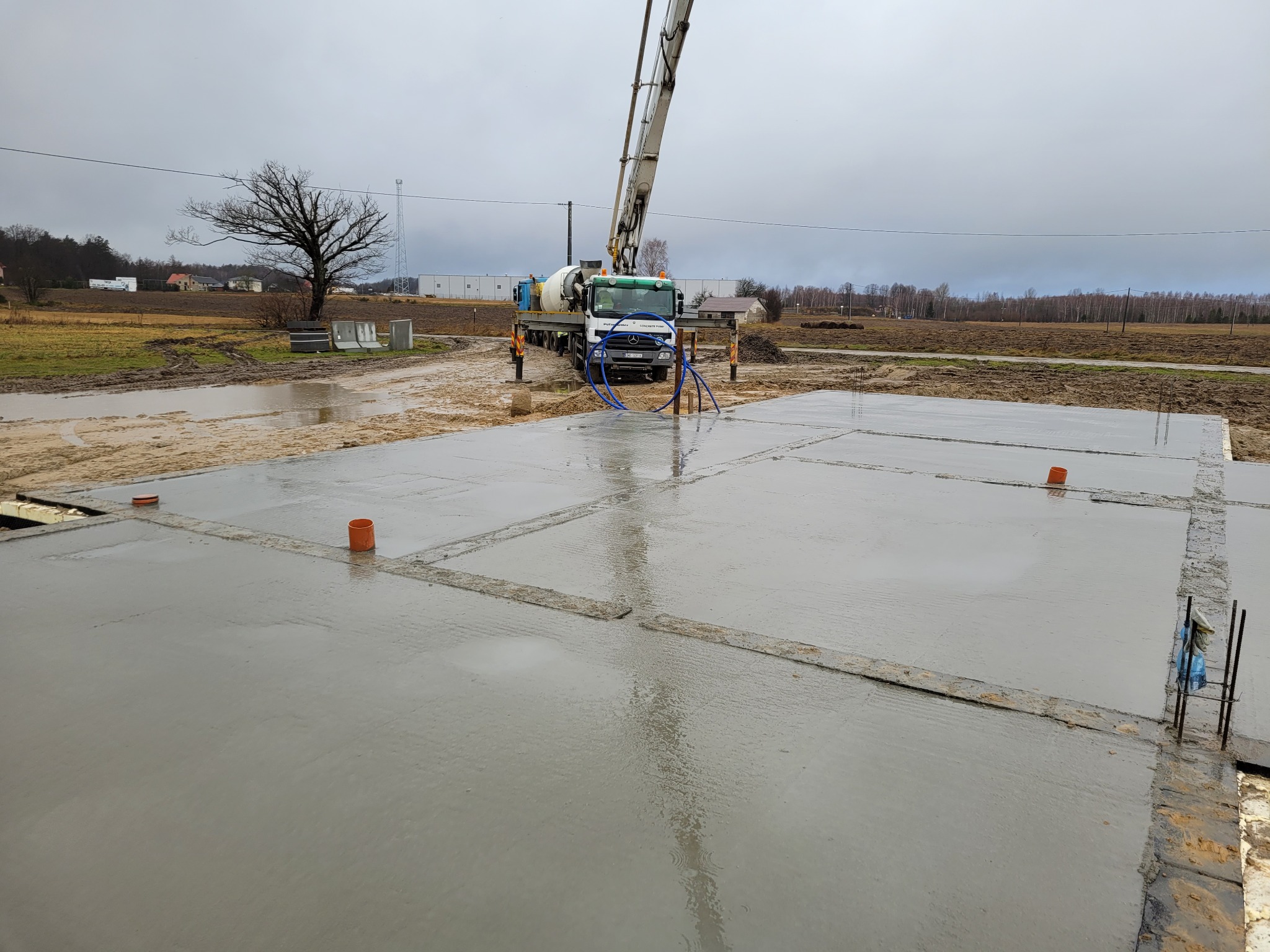 Zalany betonowy fundament z widoczną pompą do betonu i elementami szalunku. Na powierzchni betonu widoczne pomarańczowe rury.