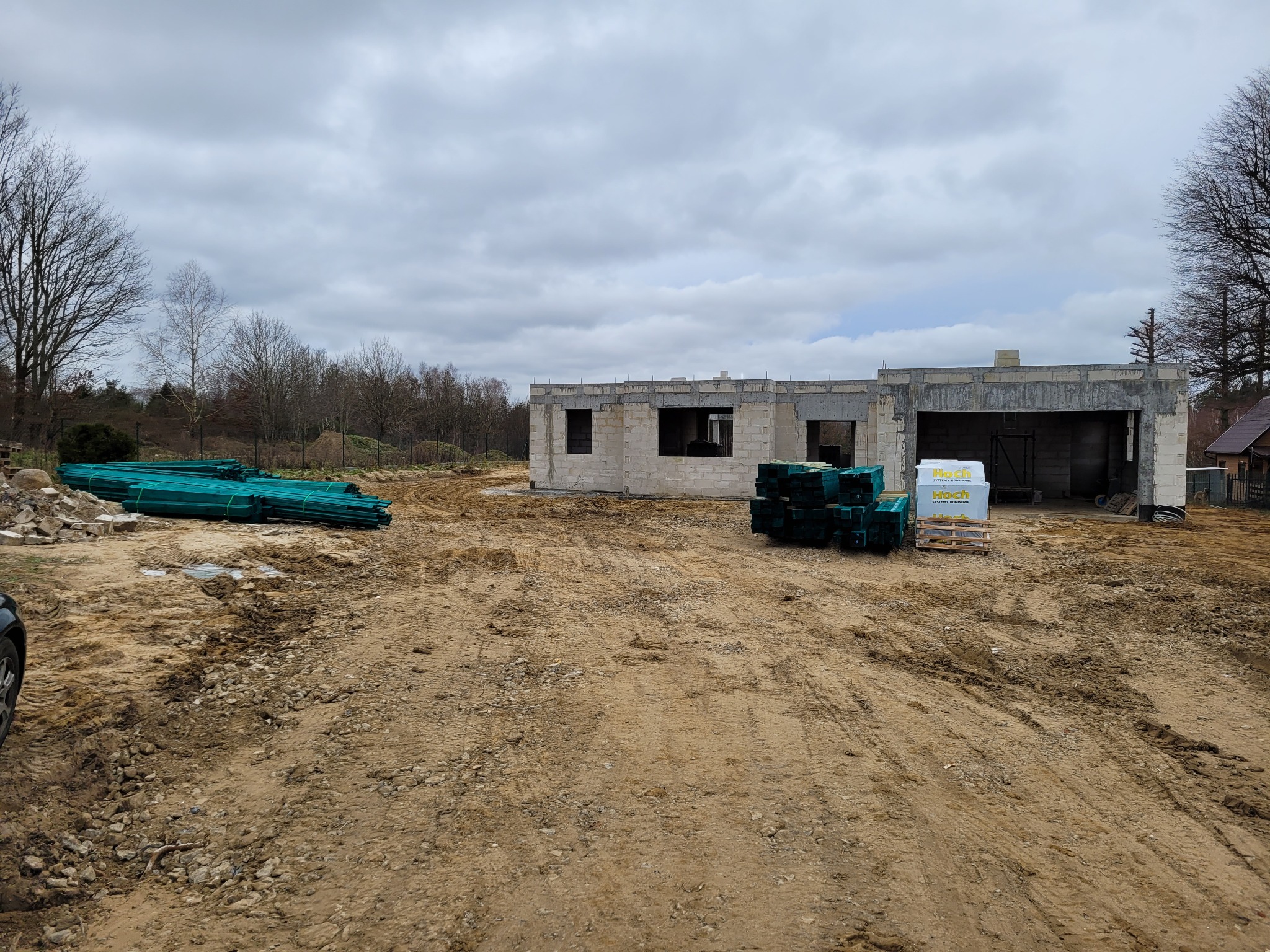 Niedokończony budynek z betonu komórkowego w stanie surowym otwartym, z widocznymi materiałami budowlanymi i ziemistym terenem wokół.