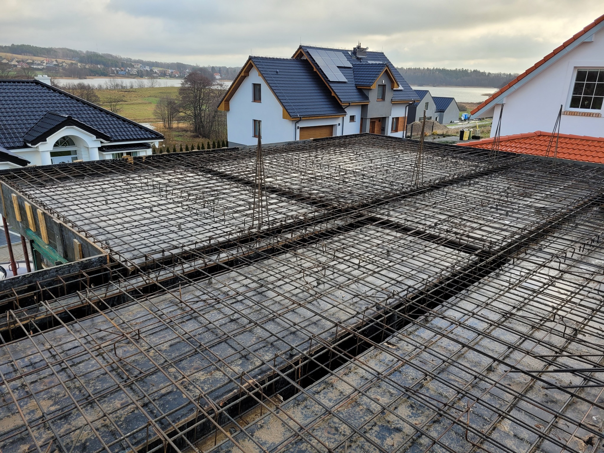 Zbrojenie stropu na budowie domu jednorodzinnego z widokiem na jezioro w tle, pochmurny dzień. Siatka z prętów stalowych ułożona na szalunku.