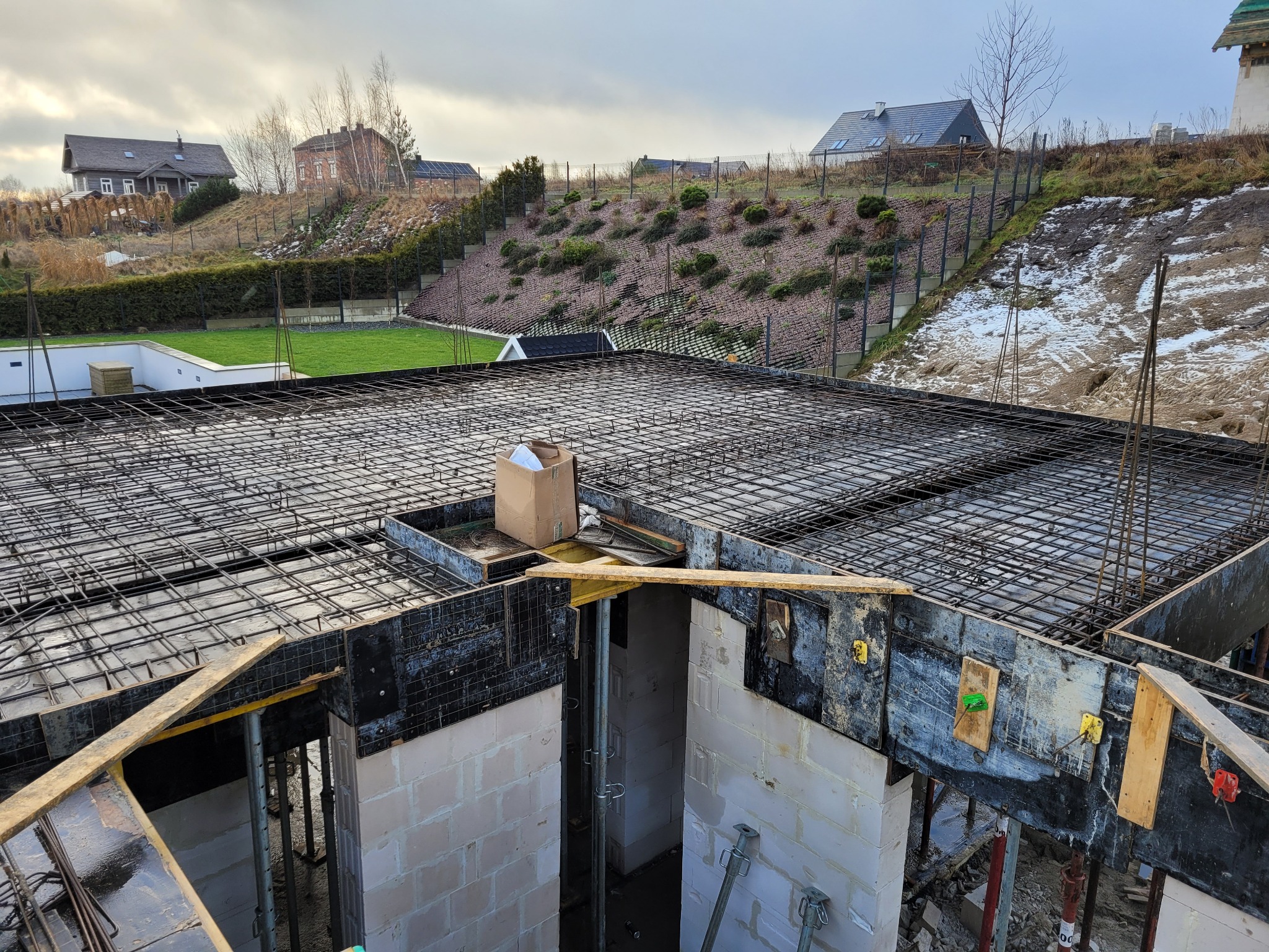 Zbrojenie stropu budynku w trakcie budowy, widoczne szalunki i podpory. W tle zabudowa jednorodzinna na wzgórzu.