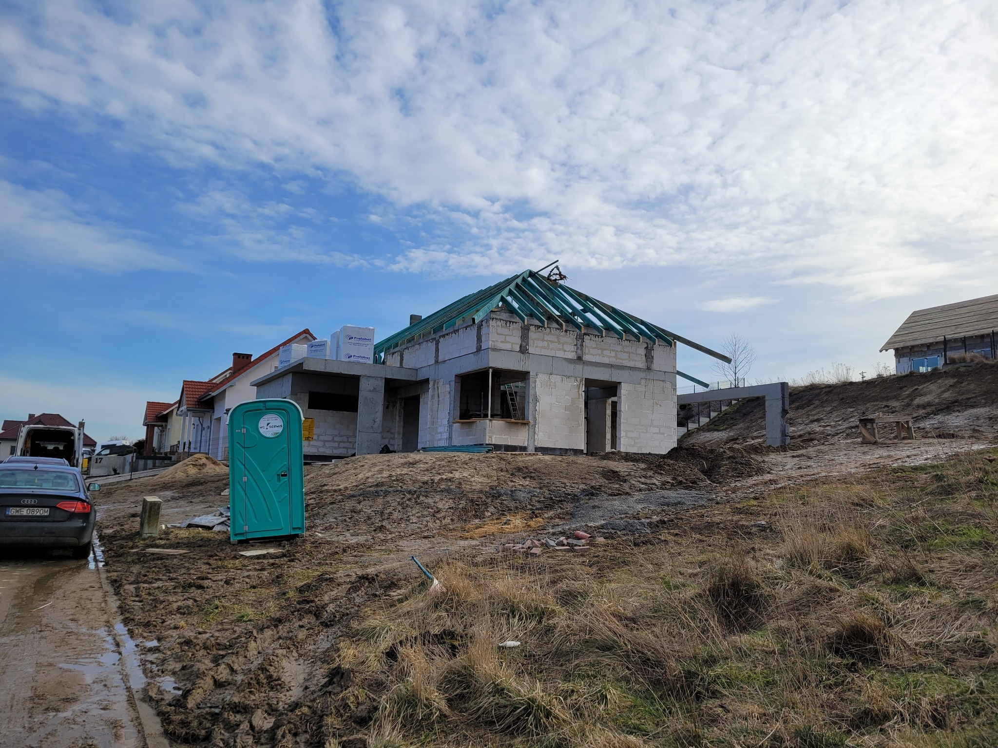 Budowa domu w trakcie realizacji: surowy budynek z bloczków, widoczna konstrukcja dachu, teren budowy, przenośna toaleta. Ujęcie z niskiej perspektywy.
