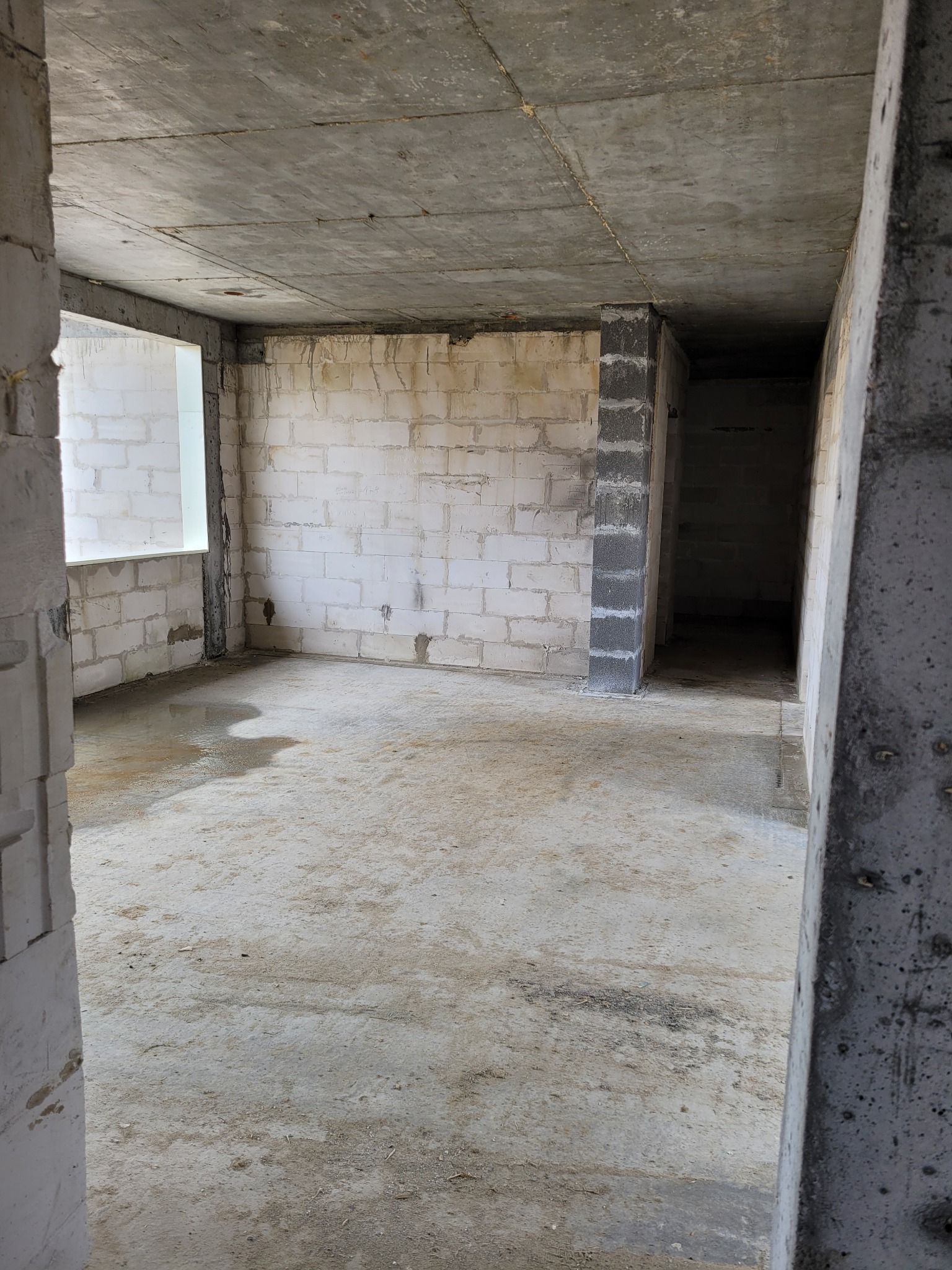 Surowe wnętrze w trakcie budowy: ściany z bloczków, betonowy sufit, widok przez otwór drzwiowy. Wnętrze w stanie deweloperskim, z widocznym oknem.