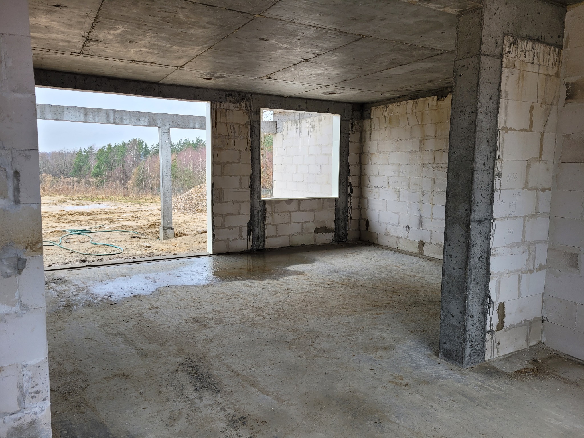 Surowy stan wnętrza domu w budowie: betonowy strop, ściany z bloczków, otwory okienne i drzwiowe z widokiem na zewnątrz. Stan surowy zamknięty.