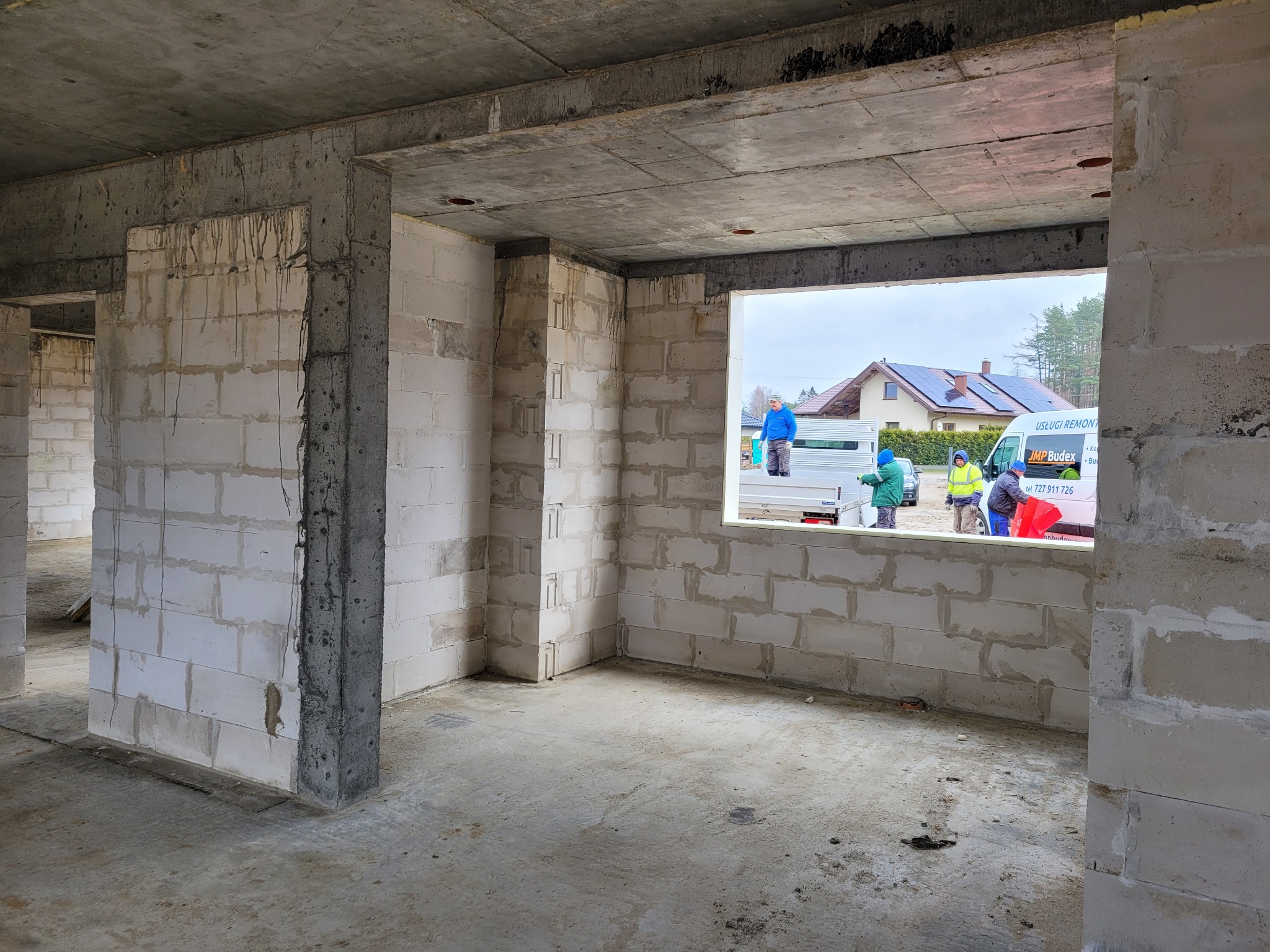 Surowy stan domu z betonu komórkowego: ściany, strop i otwór okienny. Na zewnątrz ekipa budowlana przy samochodzie. Widoczne domy z panelami słonecznymi.