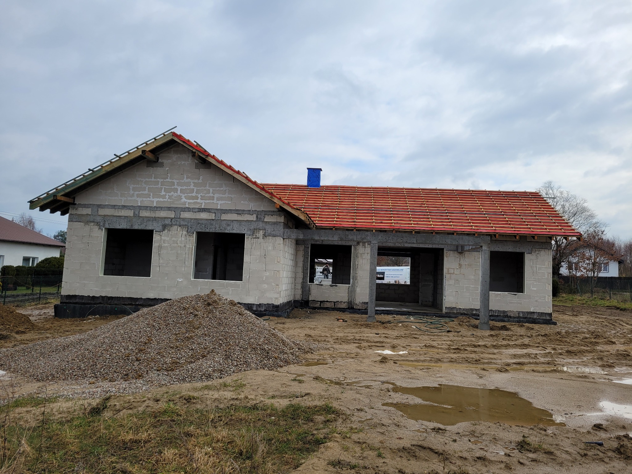 Budowa domu w trakcie: murowane ściany, częściowo położona czerwona dachówka, widoczny stos żwiru. Stan surowy otwarty, pochmurne niebo w tle.