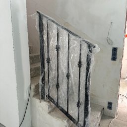 Spawpodklucz - Czarna, metalowa balustrada schodowa z ozdobnymi elementami, zabezpieczona folią ochronną podczas prac wykończeniowych na betonowych schodach w surowym wnętrzu.