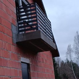 Spawpodklucz - Nowoczesna, czarna balustrada balkonowa z poziomych profili, zamontowana na surowym, betonowym balkonie w budynku z czerwonej cegły. Widok z dołu, na tle pochmurnego nieba i drzew bez liści.