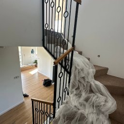 Spawpodklucz - Klatka schodowa z nową, czarną balustradą z drewnianym poręczą, częściowo zabezpieczona folią ochronną podczas remontu, widok z góry na drewnianą podłogę na dole.