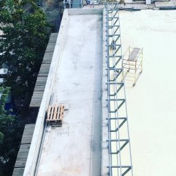 Spawpodklucz - Widok z góry na montaż metalowej konstrukcji zabezpieczającej na dachu budynku, z widocznym fragmentem miasta i drzewami w tle.