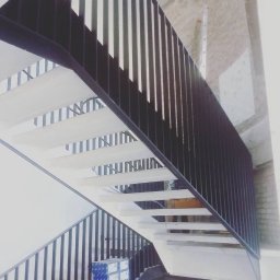 Spawpodklucz - Nowoczesne schody z białymi stopniami i czarną metalową balustradą, widok z dołu, na dole kartony z materiałami budowlanymi.