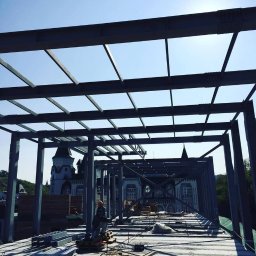 Spawpodklucz - Konstrukcja metalowa w trakcie montażu, widoczni pracownicy w kaskach, w tle budynek o architekturze pałacowej i błękitne niebo.