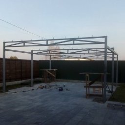 Spawpodklucz - Metalowa konstrukcja z profili zamkniętych w trakcie budowy z widocznymi narzędziami i elementami montażowymi na kostce brukowej, na tle drewnianego i metalowego ogrodzenia.