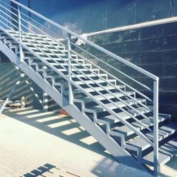 Spawpodklucz - Zewnętrzne schody metalowe z ażurowymi stopniami i prostą balustradą, pomalowane na jasnoszary kolor, prowadzące do budynku z ciemną, kafelkową elewacją.