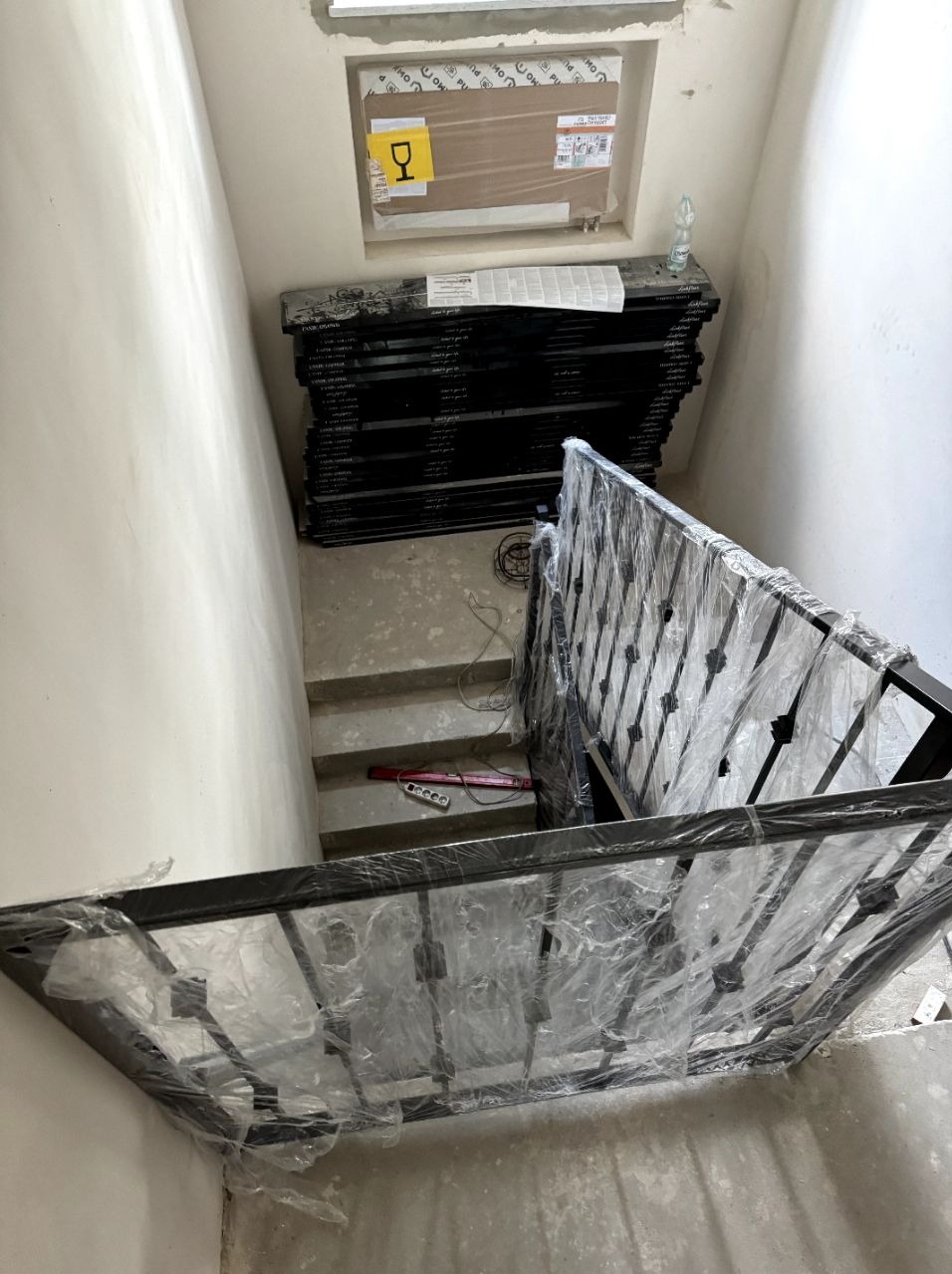 Częściowo zamontowana czarna metalowa balustrada schodowa zabezpieczona folią ochronną, z pakietami elementów balustrady składowanymi na podeście schodowym, widok z góry na surowy stan wykończenia...