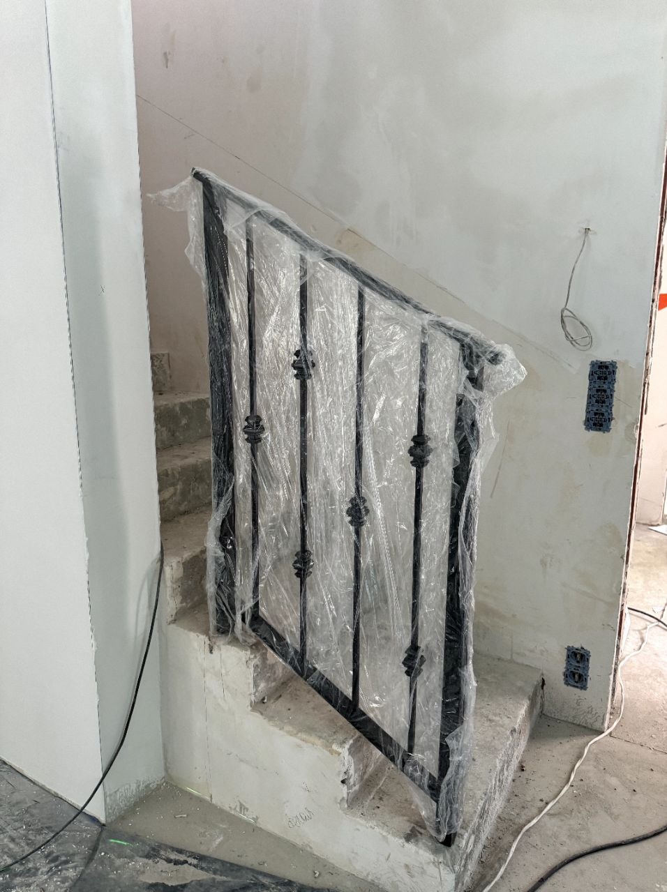 Czarna, metalowa balustrada schodowa z ozdobnymi elementami, zabezpieczona folią ochronną podczas prac wykończeniowych na betonowych schodach w surowym wnętrzu.