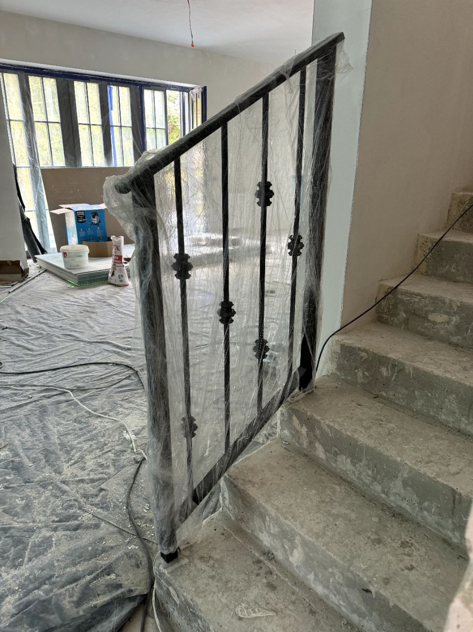 Czarne metalowe balustrady schodowe z dekoracyjnymi elementami, zabezpieczone folią ochronną podczas remontu, widoczne surowe betonowe schody i zabezpieczona folią podłoga.