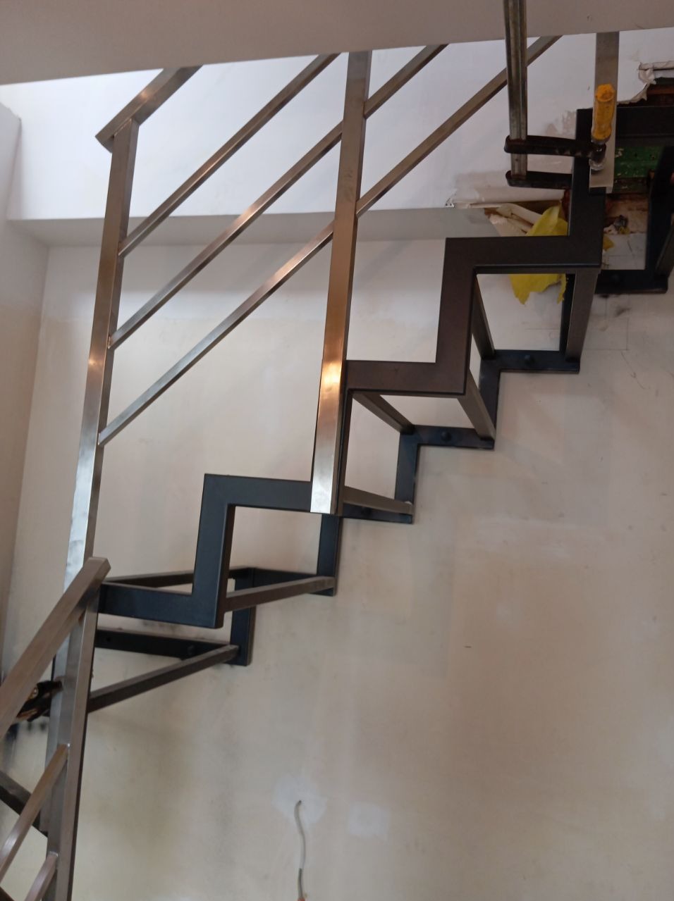 Metalowe schody w trakcie montażu, z widoczną konstrukcją stopni i częścią balustrady, przymocowane do ściany z widocznymi śladami po instalacji i narzędziem monterskim u góry.
