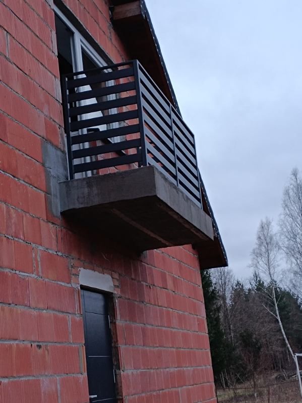 Nowoczesna, czarna balustrada balkonowa z poziomych profili, zamontowana na surowym, betonowym balkonie w budynku z czerwonej cegły. Widok z dołu, na tle pochmurnego nieba i drzew bez liści.