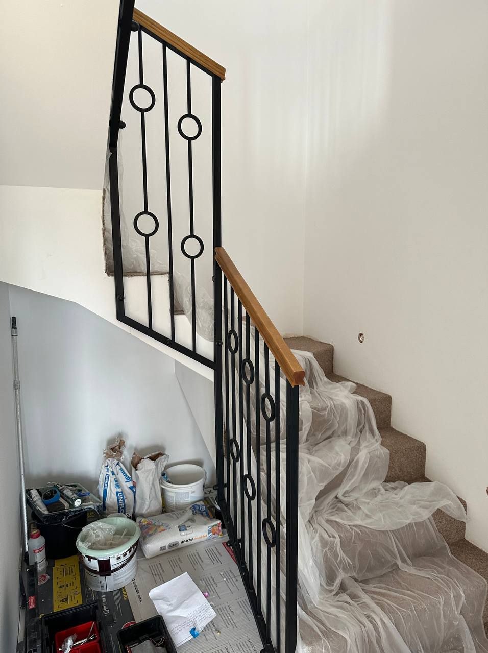 Czarna, metalowa balustrada schodowa z drewnianym poręczą, z okrągłymi elementami dekoracyjnymi, zamontowana w jasnym wnętrzu podczas remontu, schody zabezpieczone folią ochronną, widoczne...