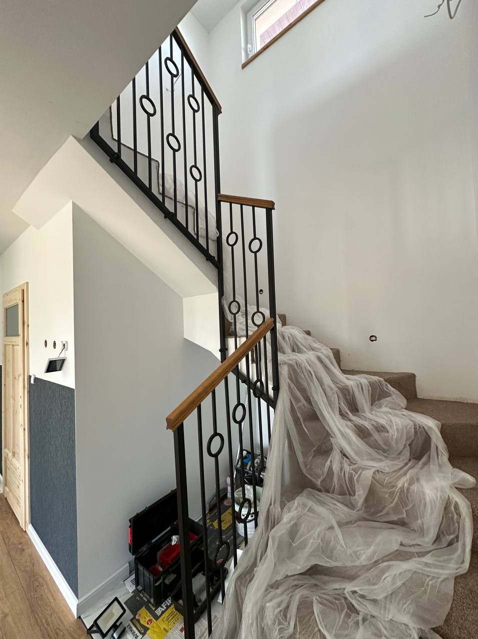 Wnętrze domu w trakcie prac wykończeniowych: schody z czarną metalową balustradą z drewnianym poręczem, zabezpieczone folią malarską, obok skrzynka z narzędziami.