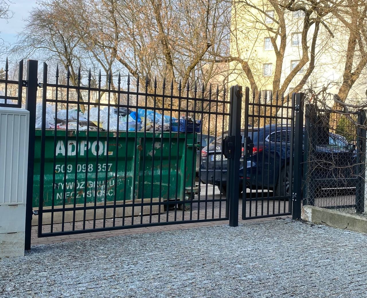 Czarne metalowe ogrodzenie z bramą wjazdową, za którym widać kontener na śmieci i zaparkowany samochód osobowy.