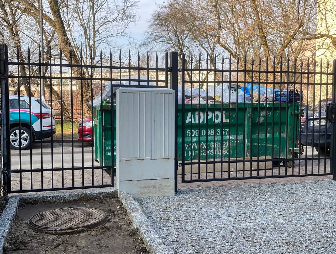 Czarna metalowa brama z ostrymi zakończeniami prętów, widok na ulicę z kontenerem na śmieci w tle, studzienka kanalizacyjna w ziemi przed bramą.