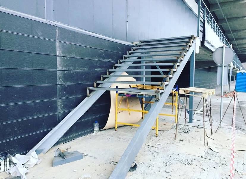 Konstrukcja metalowych schodów podczas montażu w surowym wnętrzu, widoczne rusztowania, narzędzia i zabezpieczenia budowlane.