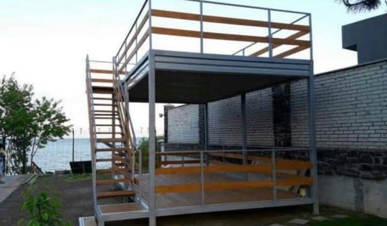 Dwupoziomowa konstrukcja metalowa z drewnianymi elementami, schodami i balustradami, z widokiem na morze w tle.