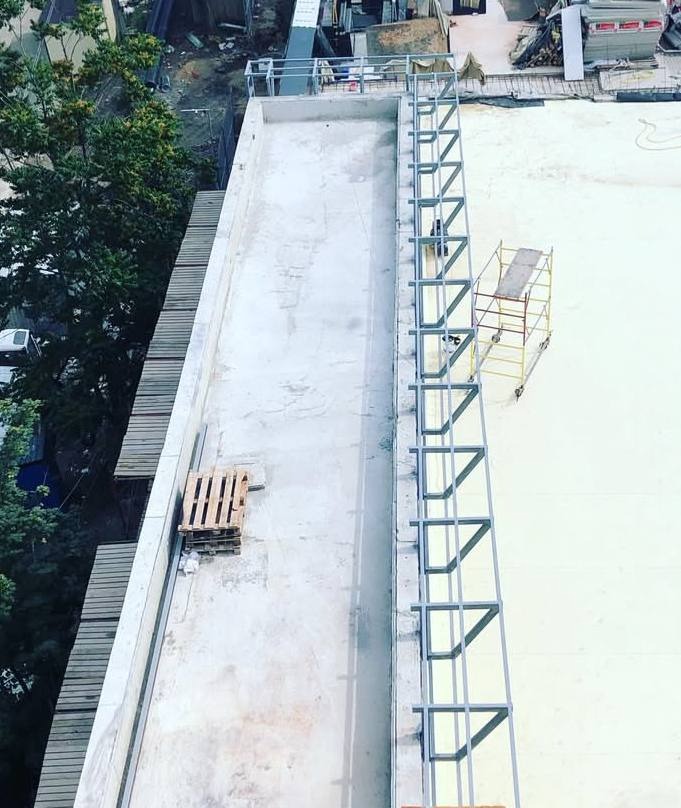 Widok z góry na montaż metalowej konstrukcji zabezpieczającej na dachu budynku, z widocznym fragmentem miasta i drzewami w tle.