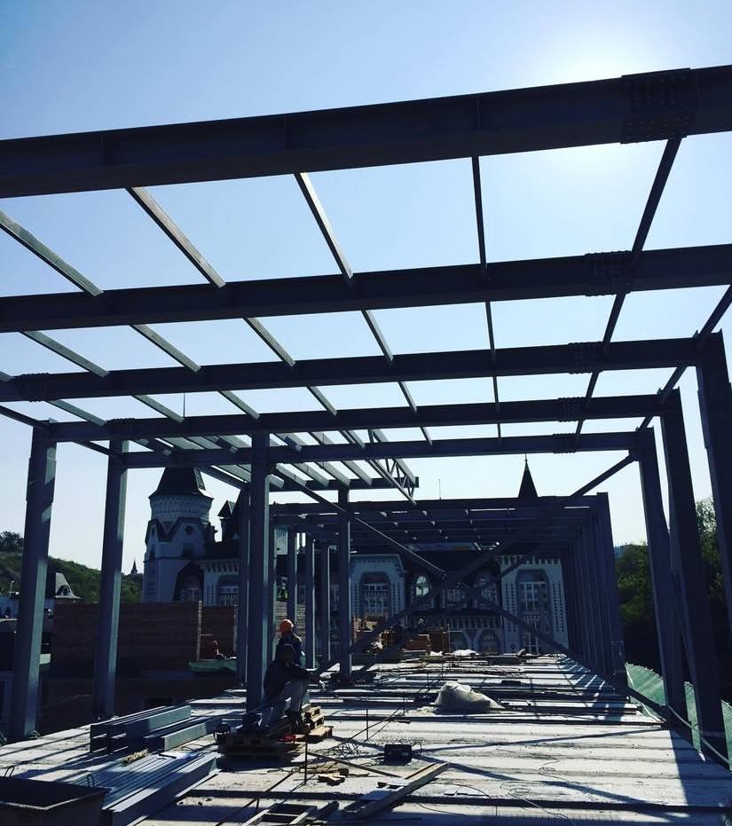 Konstrukcja metalowa w trakcie montażu, widoczni pracownicy w kaskach, w tle budynek o architekturze pałacowej i błękitne niebo.