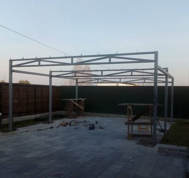 Metalowa konstrukcja z profili zamkniętych w trakcie budowy z widocznymi narzędziami i elementami montażowymi na kostce brukowej, na tle drewnianego i metalowego ogrodzenia.