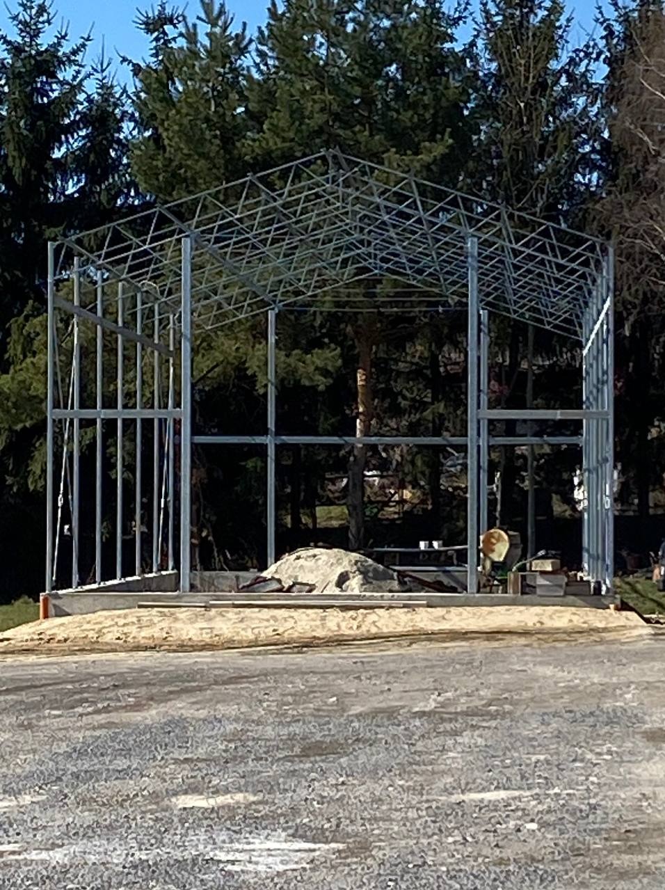 Konstrukcja metalowa szkieletu budynku z widocznymi elementami zespawanymi, w tle drzewa iglaste i niebo.