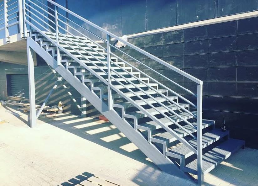 Zewnętrzne schody metalowe z ażurowymi stopniami i prostą balustradą, pomalowane na jasnoszary kolor, prowadzące do budynku z ciemną, kafelkową elewacją.