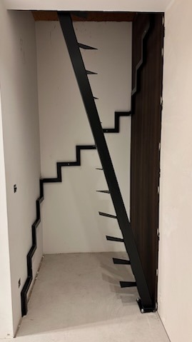 Surowe, metalowe schody modułowe w trakcie montażu w wąskim, niedokończonym wnętrzu, z widocznymi elementami mocującymi do ściany i pionową podporą, bez poręczy.