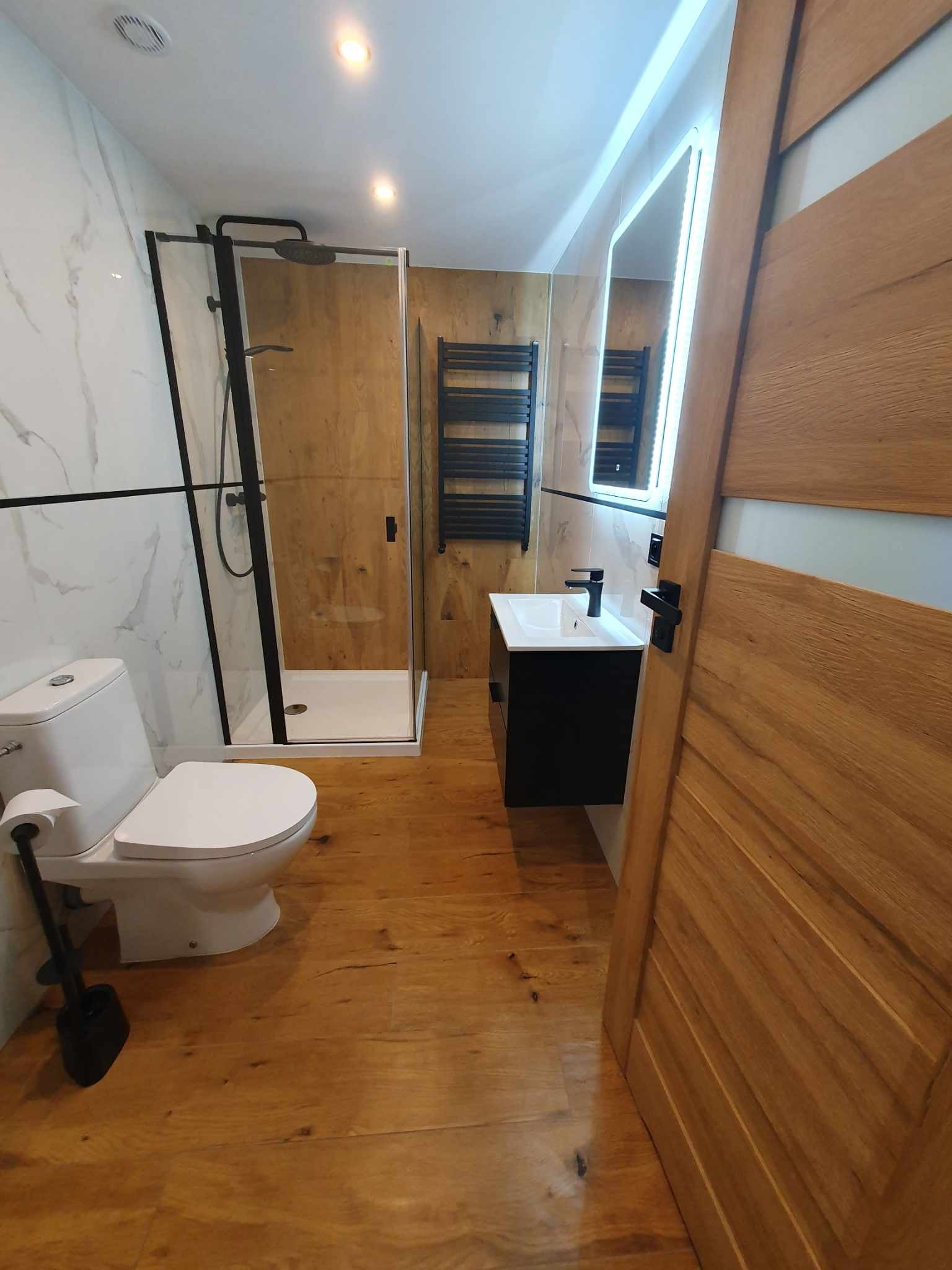 Nowoczesna łazienka z kabiną prysznicową typu walk-in z czarnymi ramami, umywalką na szafce w kolorze czarnym, podświetlanym lustrem LED i drewnianą podłogą oraz częściowo drewnianą ścianą.