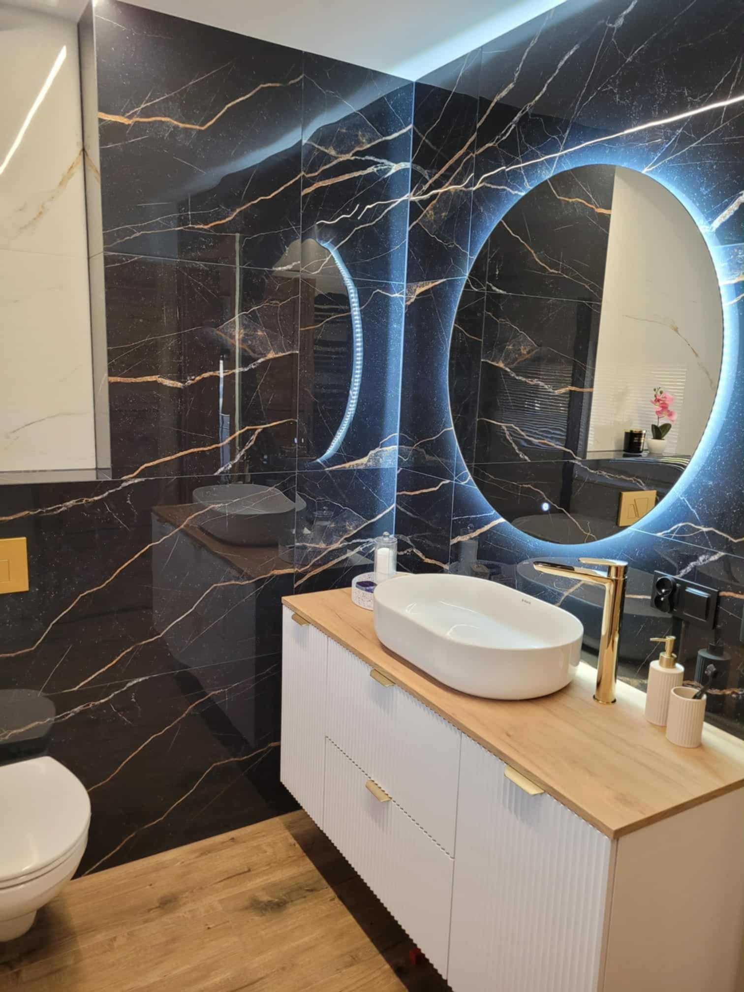 Elegancka łazienka z czarnymi płytkami imitującymi marmur, podświetlanym lustrem i białą szafką z drewnianym blatem. Złota armatura i drewniana podłoga dodają luksusu.