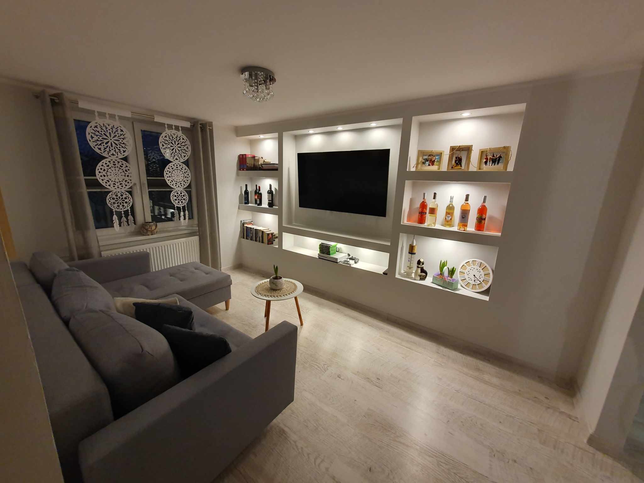 Salon z szarą sofą i ścianą z wnękami podświetlonymi LED, w których stoją książki, dekoracje i butelki. Na ścianie wisi telewizor. Drewniana podłoga.