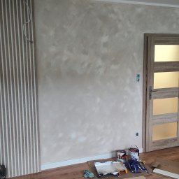 FIRMA OGÓLNOBUDOWLANA EMILIAN LISIEWICZ - Ściana z efektem dekoracyjnym w beżach, listwy ścienne, drzwi z przeszkleniami. Na podłodze akcesoria malarskie i kartony. Widoczne gniazdka elektryczne.