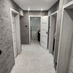 FIRMA OGÓLNOBUDOWLANA EMILIAN LISIEWICZ - Wykończony korytarz z marmurową podłogą i dekoracyjną szarą ścianą. Widoczne trzy białe drzwi z czarnymi klamkami i oświetlenie sufitowe.