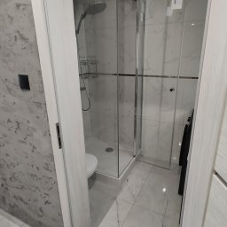 FIRMA OGÓLNOBUDOWLANA EMILIAN LISIEWICZ - Wykończona łazienka z kabiną prysznicową typu walk-in, białe marmurowe płytki na ścianach i podłodze, widok zza otwartych drzwi, nowoczesny design.