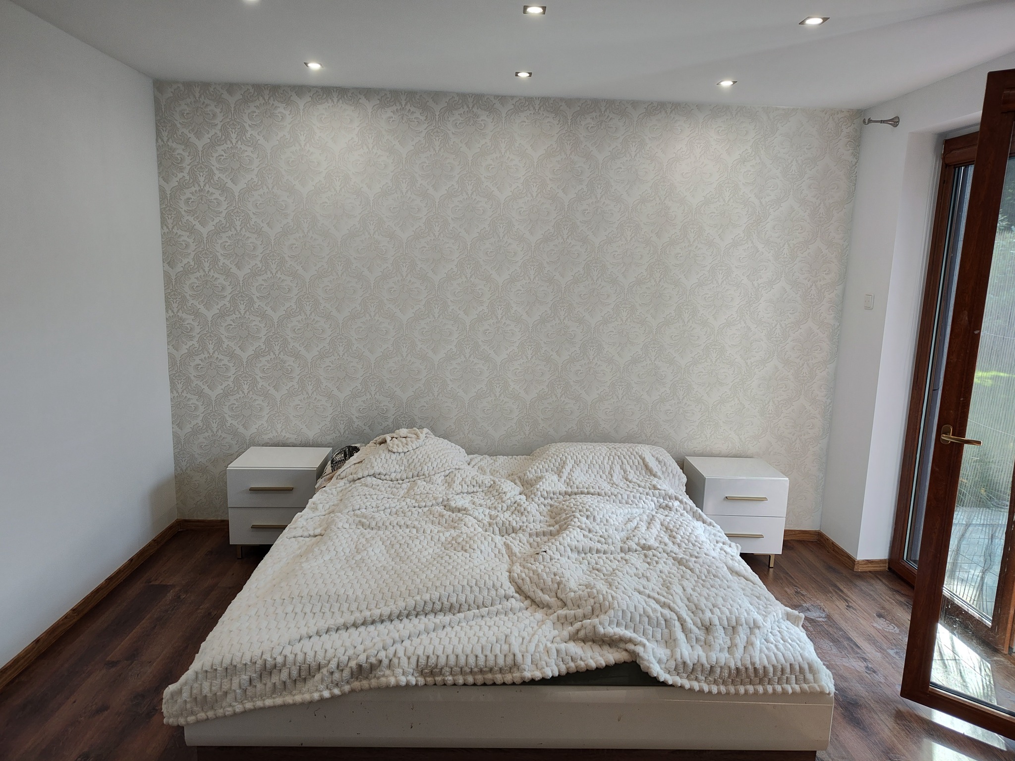 Sypialnia po remoncie: łóżko z białą, teksturowaną narzutą, białe szafki nocne z złotymi uchwytami, ściana w jasne, wzorzyste tapety, drewniana podłoga i drzwi balkonowe.