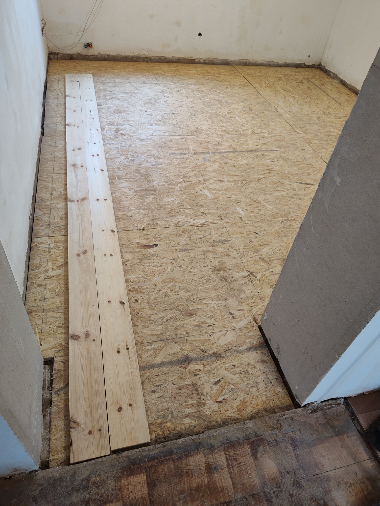 Przygotowanie podłogi z płyty OSB w trakcie remontu, widoczne deski podłogowe czekające na montaż, surowe ściany i wystające kable.