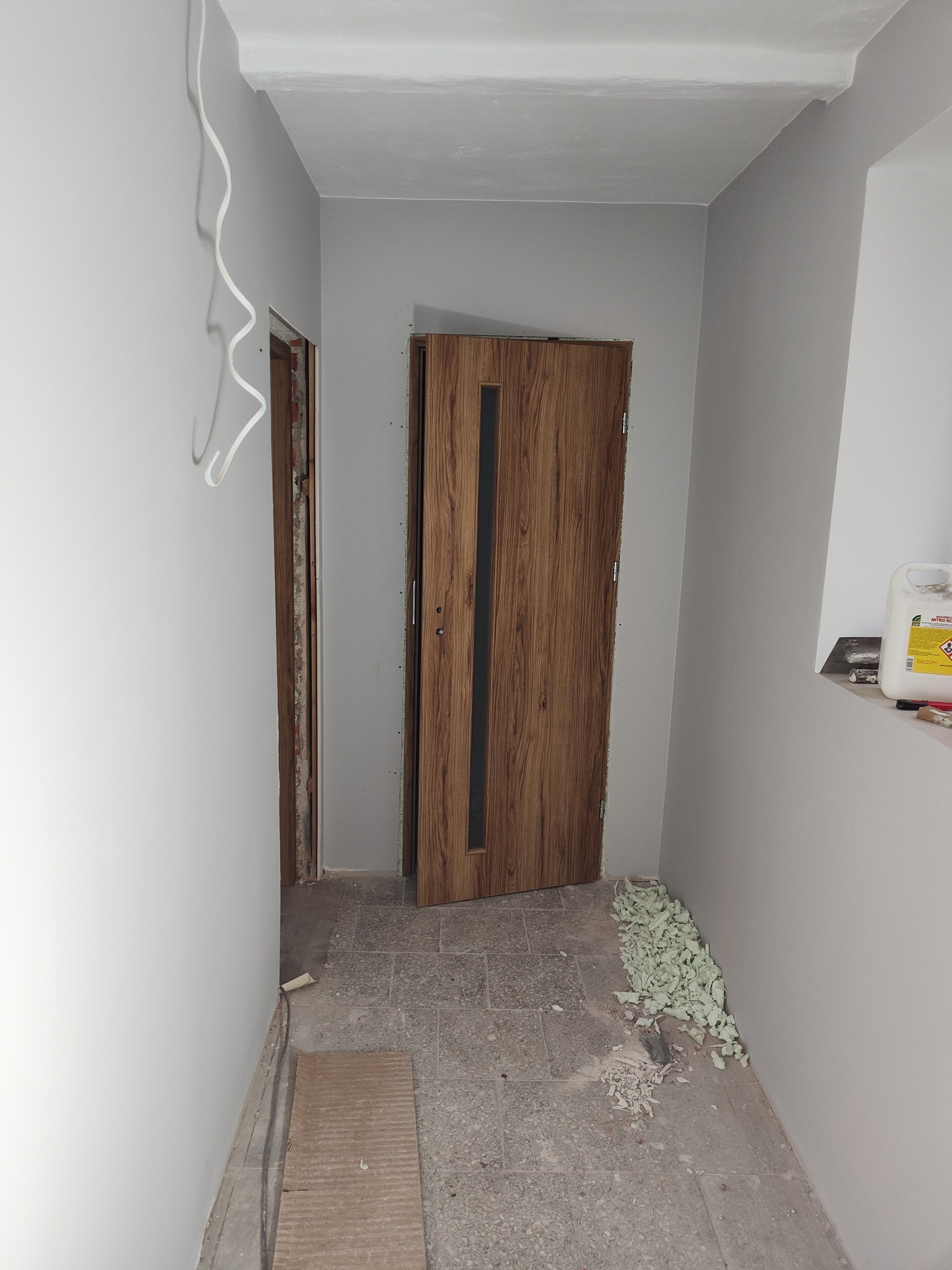 Korytarz w trakcie prac wykończeniowych: świeżo pomalowane szare ściany, osadzona drewniana futryna i drzwi z czarnym pasem, widoczne przewody elektryczne i resztki pianki montażowej na podłodze.