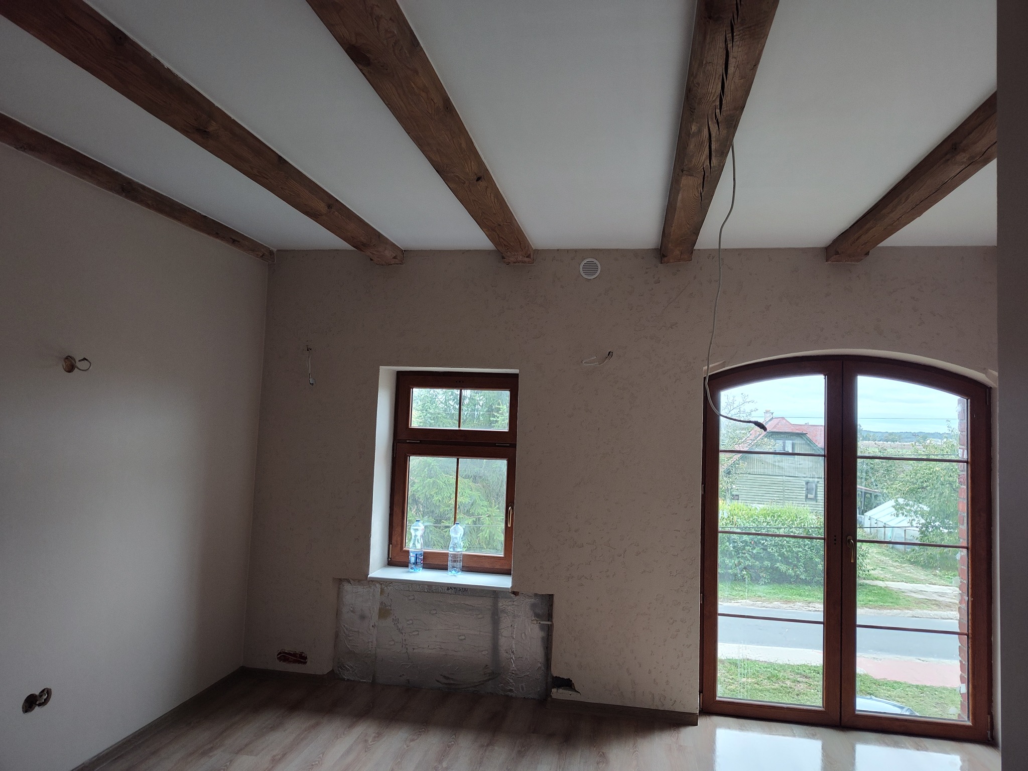 Wnętrze pokoju po remoncie z drewnianymi belkami stropowymi, drewnianą stolarką okienną, częściowo zabezpieczonym grzejnikiem i gładzią na ścianach.