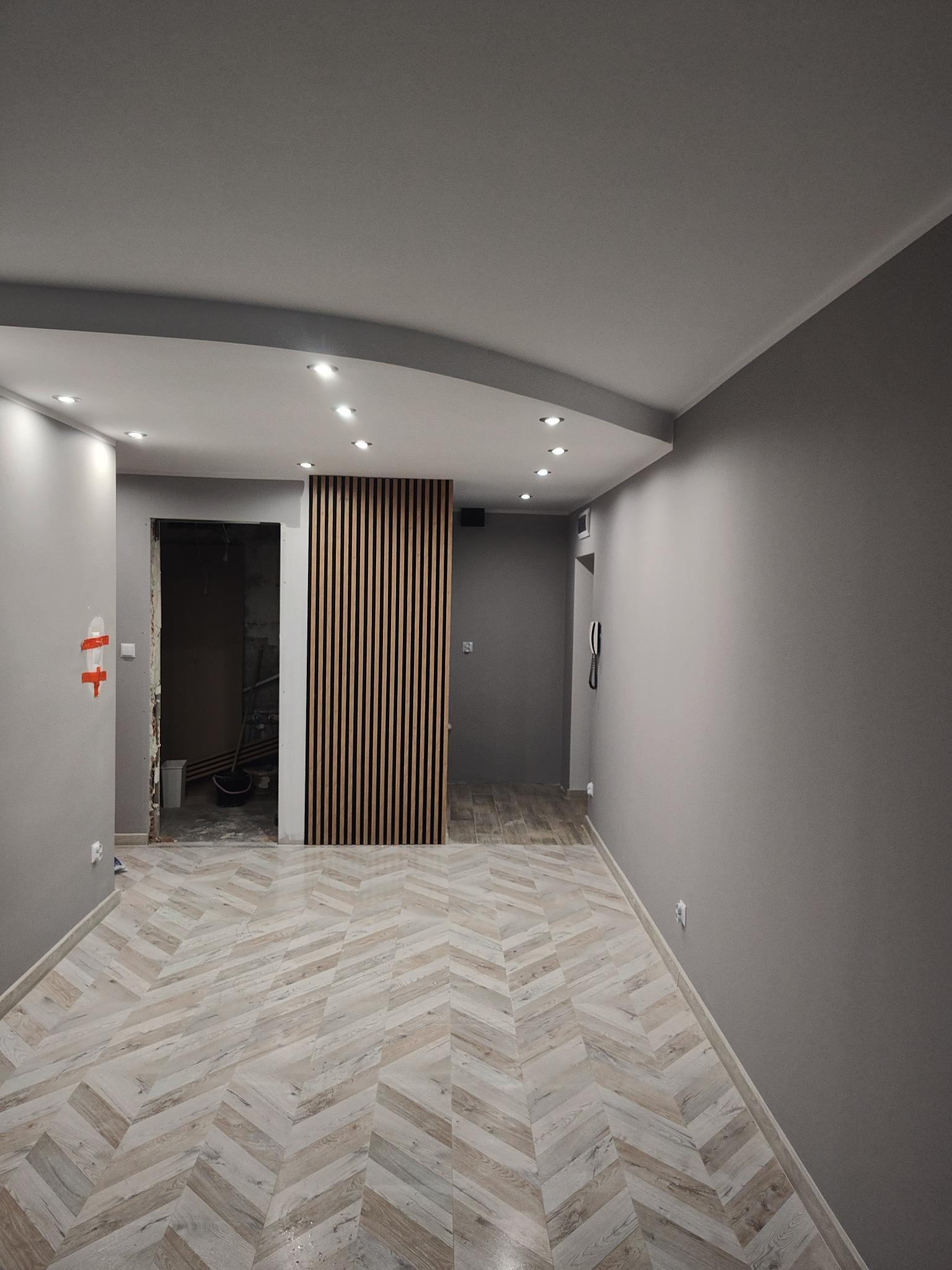 Wnętrze mieszkania w trakcie remontu: ułożona podłoga w jodełkę, drewniane lamele na ścianie, widoczne przygotowania do remontu łazienki z pozostawionym wiadrem i narzędziami.