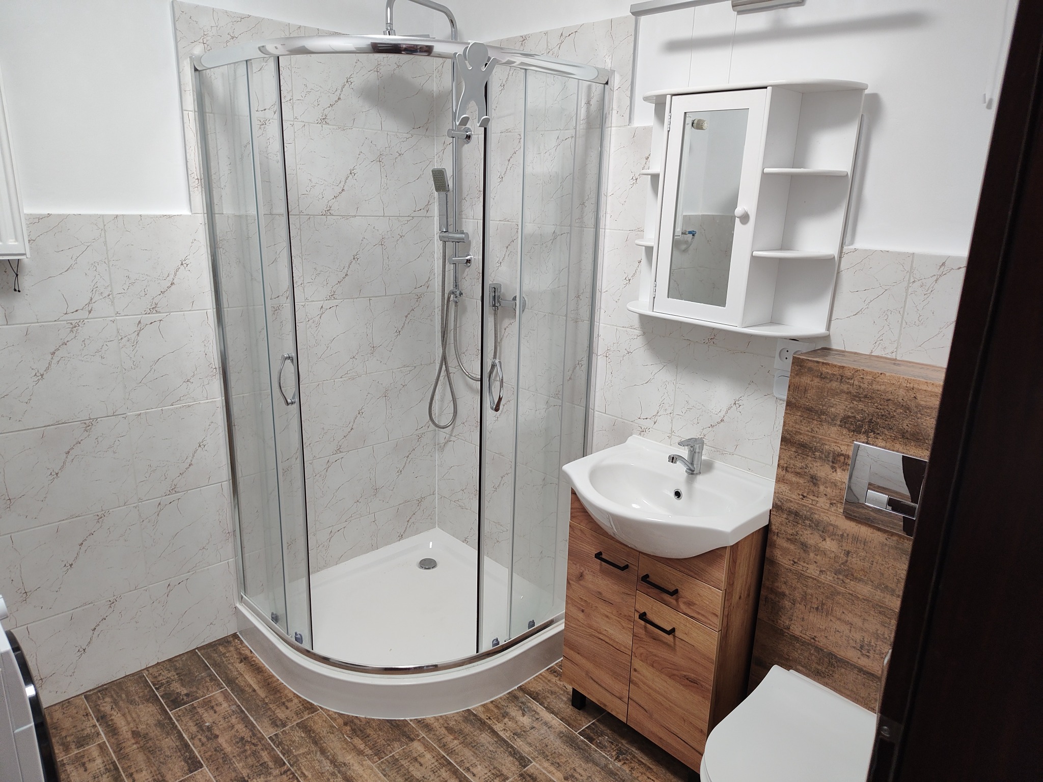 Nowoczesna łazienka z kabiną prysznicową narożną, umywalką na szafce z drewnopodobnym frontem i wiszącą szafką z lustrem, wykończona płytkami imitującymi marmur i drewnem.