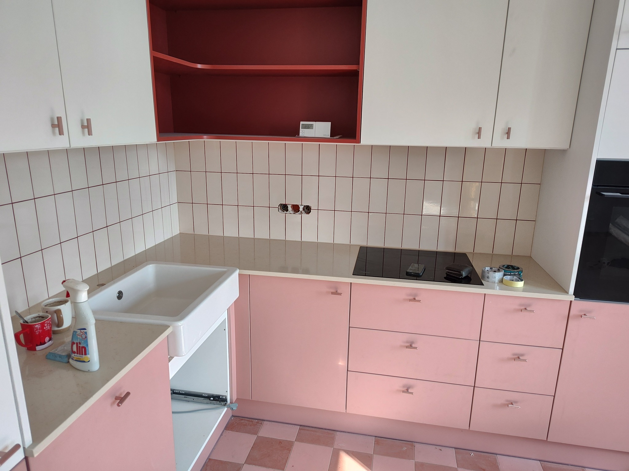 Fragment odnowionej kuchni z różowymi szafkami, białym zlewem farmerskim i białymi płytkami ściennymi z różową fugą. Na blacie widoczne akcesoria i środki czystości.